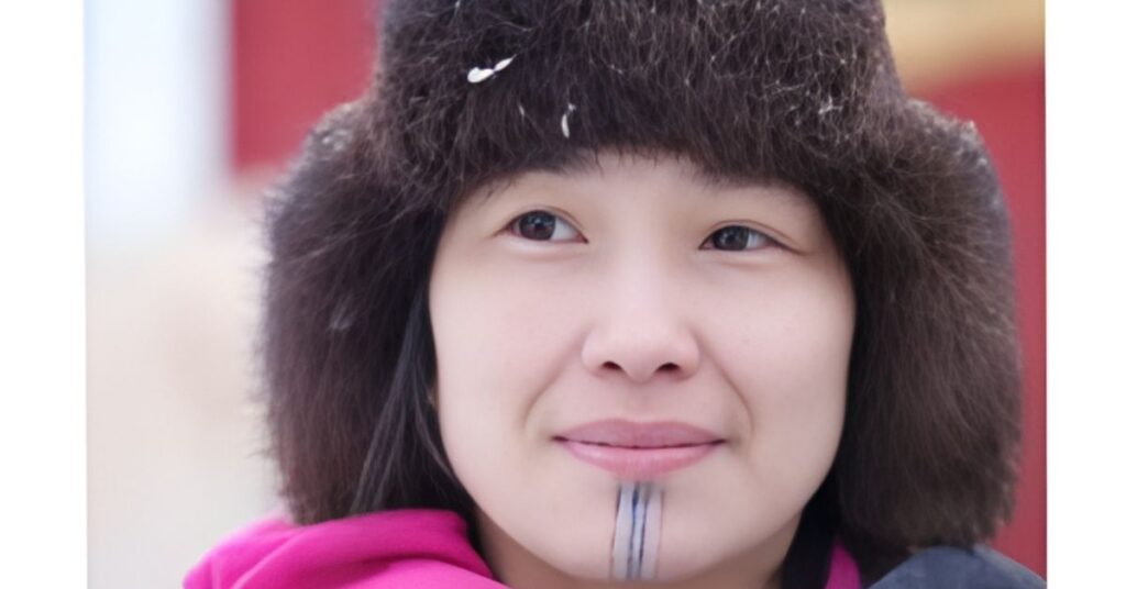 A close-up shot of a person wearing a fur hat with snow on it and a pink jacket. They have a tattoo on their chin.