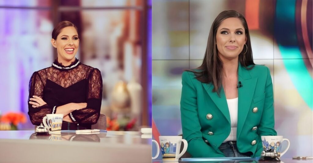 A person in a black lace dress sits behind a desk with arms crossed and smiles.