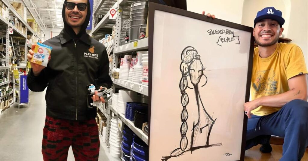 A person in a yellow t-shirt and blue sweatpants holds a framed drawing of a stylized figure with long braided hair. The drawing is in black ink on a white background and signed at the bottom righ