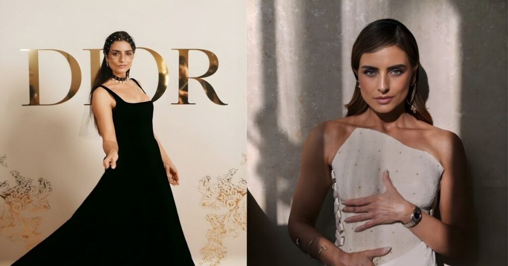 A person poses with a hand on their chest and the other on their stomach, wearing a light-colored, asymmetrical top with lace-up detailing and a white skirt.