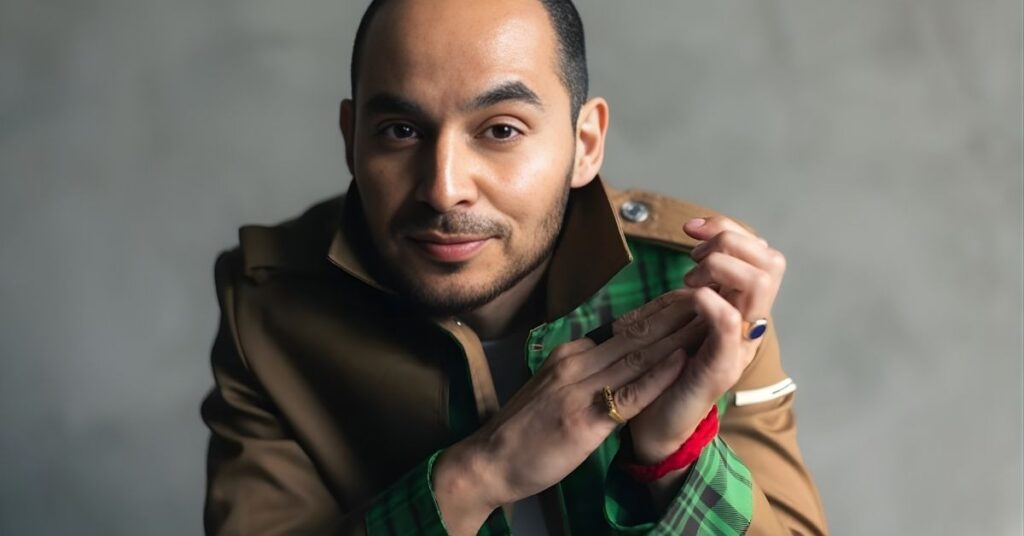 A person wearing a tan jacket with green plaid lining smiles and clasps their hands in front of them. They wear rings on their fingers and a red bracelet on their wrist.