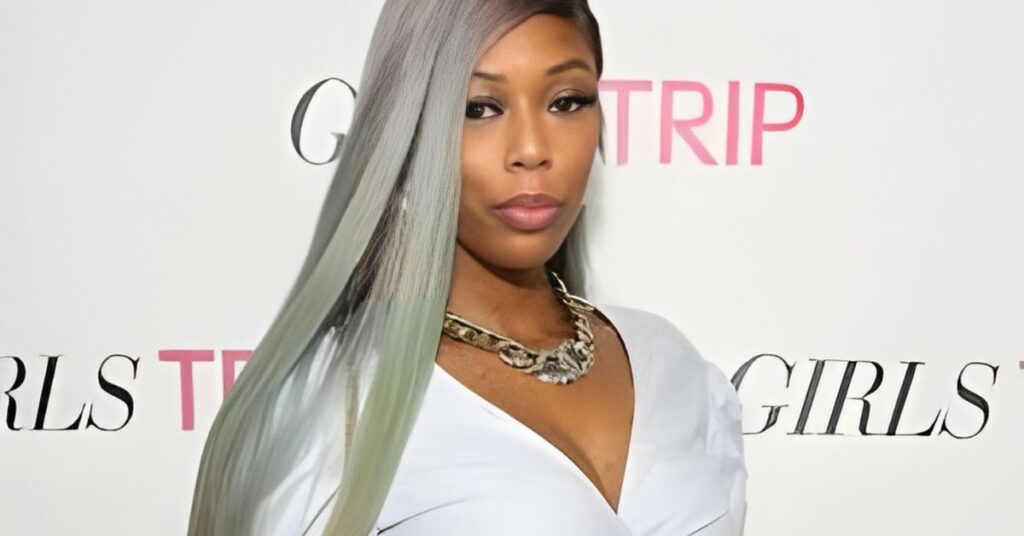 A person with long, grey-purple hair and a silver chain necklace wearing a light blue top in front of a white backdrop with the words GIRLS TRIP visible.