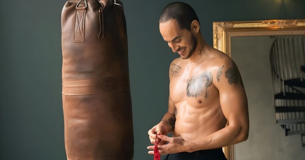 A shirtless person with tattoos stands next to a brown leather punching bag, tying a red ribbon.