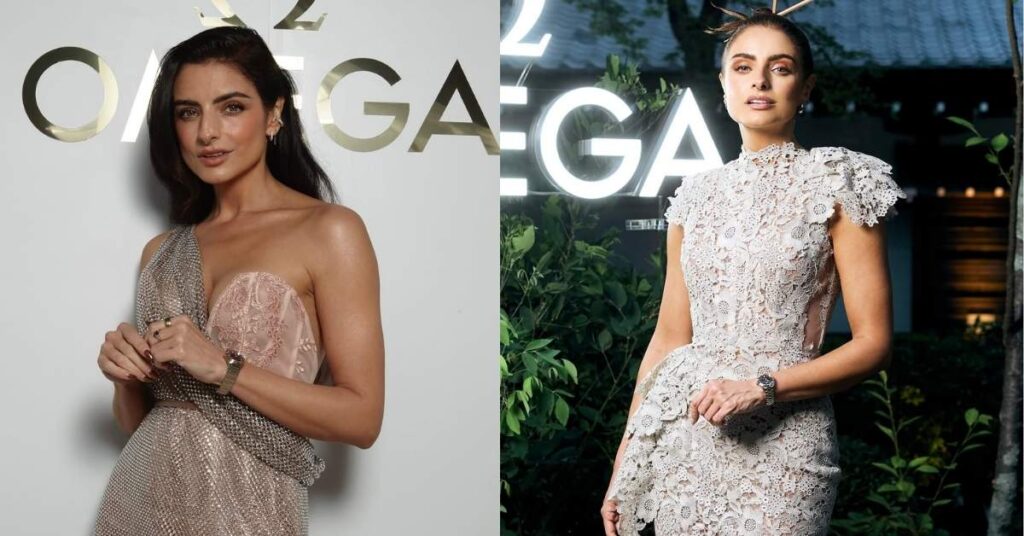 A woman in a shimmering, one-shoulder dress stands in front of an Omega logo.