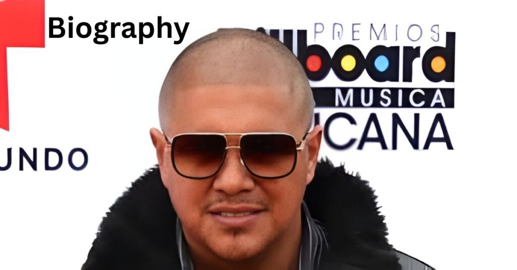 Amado Vargas with a shaved head and sunglasses wears a black coat with a fur collar. He stands in front of a white backdrop with logos that read PREMIOS board MUSICA and UNDO.