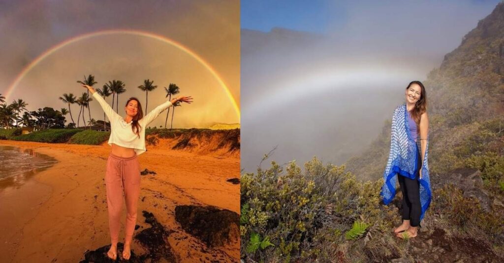 Amanda Eller stands with arms outstretched on a sandy beach, with a vibrant rainbow arching across a cloudy sky behind them. Palm trees and lush greenery line the shore.