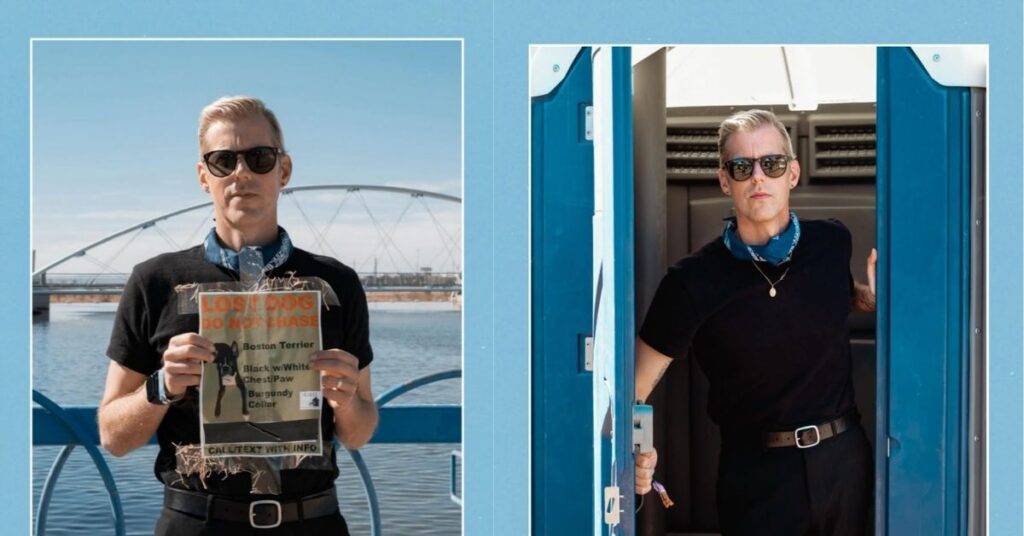 Andrew McMahon, wearing sunglasses, a black t-shirt, and black pants, stands in the doorway of a portable toilet.