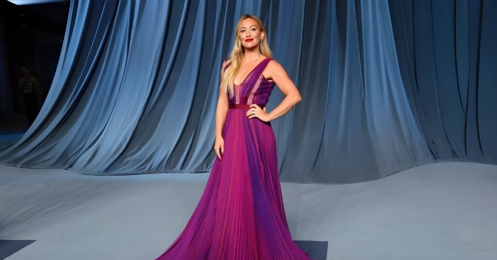Hilary Duff poses in a flowing, pleated gown that transitions from deep purple to magenta, with a V-neckline and a matching belt.