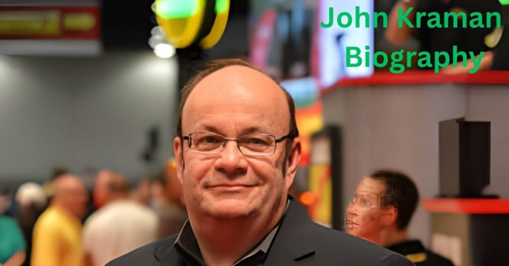John Kraman, wearing glasses and a dark jacket, smiles at the camera, with a blurred background of people and displays.