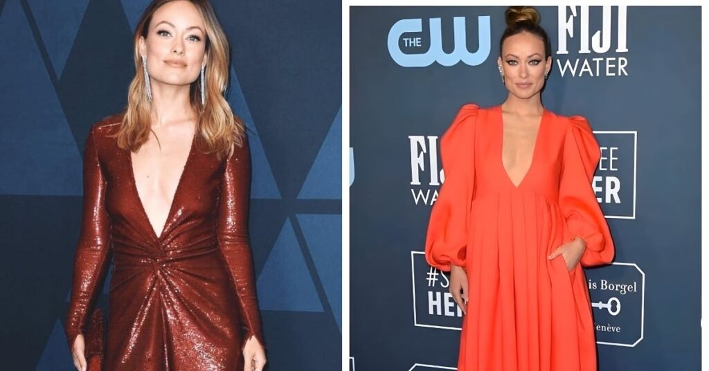 Olivia Wilde, wearing a bright orange V-neck dress with puffed sleeves and silver shoes, stands on a blue carpet in front of a backdrop with various logos.