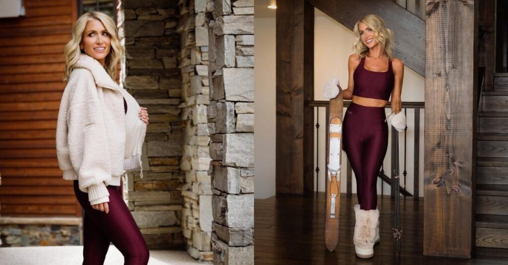 Kalee Rogers, wearing a burgundy athletic outfit and fur-lined boots, stands indoors next to a wooden staircase, holding a vintage wooden ski and poles.
