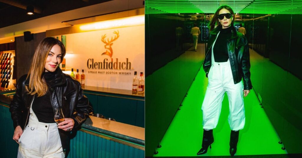 Michelle Monaghan stands at a bar with a Glenfiddich logo behind them and a glass of whisky in their hand.