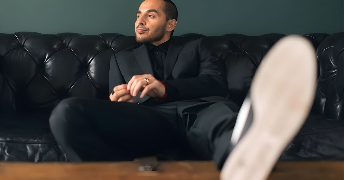 A person in a black suit sits on a black leather couch with their legs crossed, looking to the side.