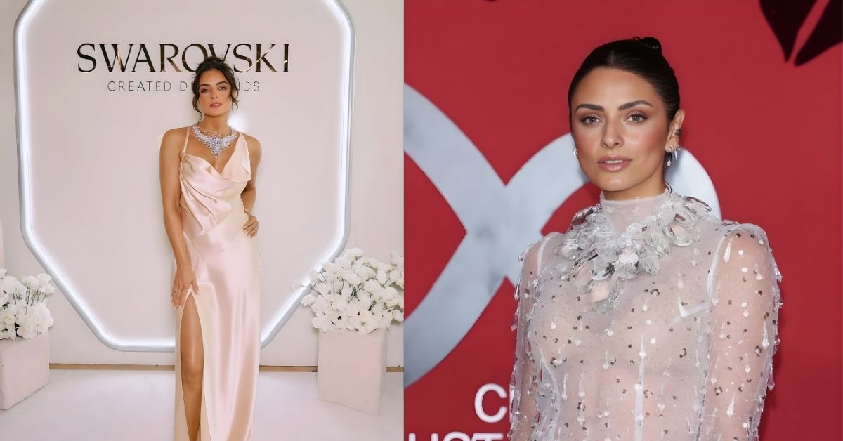 A person wearing a sheer, sparkling dress with a high neckline and intricate crystal embellishments stands against a red backdrop with a large silver XX logo.