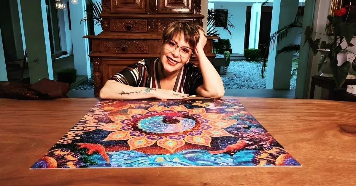 A person wearing glasses and a striped shirt leans on a table with a colorful, completed jigsaw puzzle featuring a mandala design and fantastical creatures.