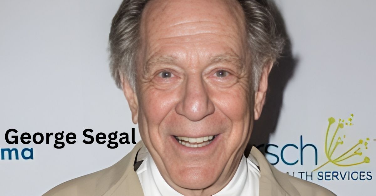A smiling person in a tan jacket and red tie stands in front of a backdrop with logos.