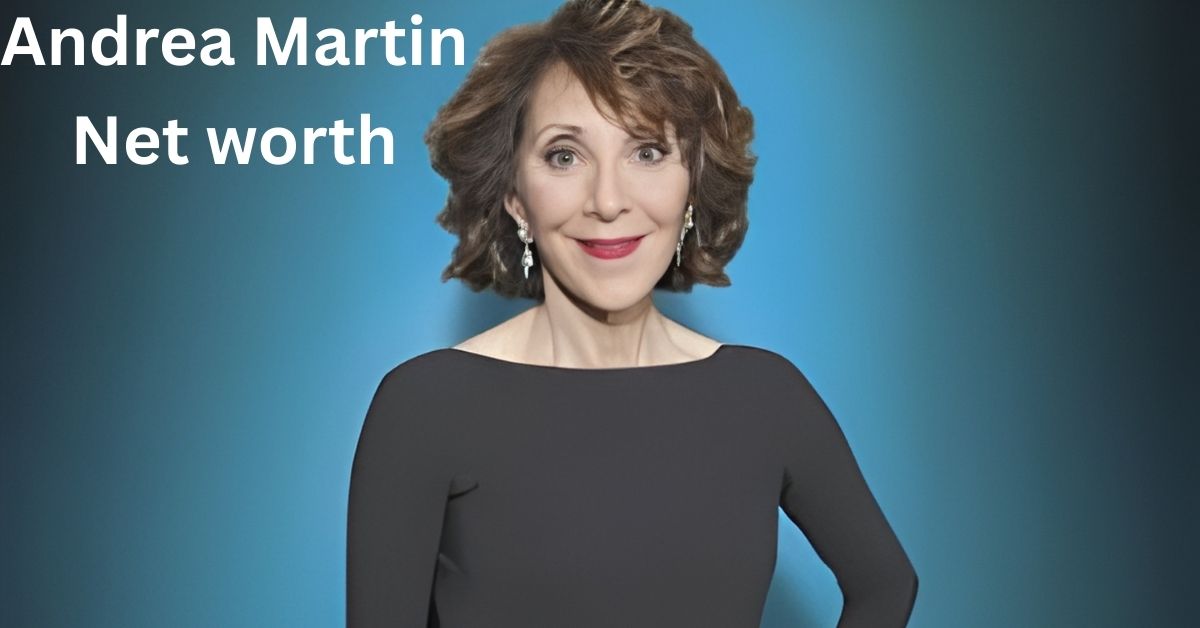 Andrea Martin, with short, brown, curly hair and red lipstick, smiles at the camera. She is wearing a black, off-the-shoulder top and dangling earrings.