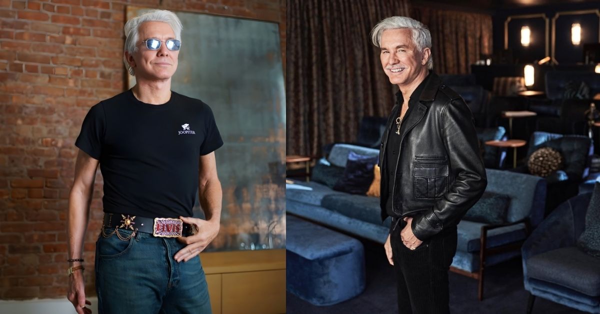 Baz Luhrmann, with gray hair and a mustache, wears a black leather jacket and black pants, standing in a room with blue velvet seating and dim lighting.
