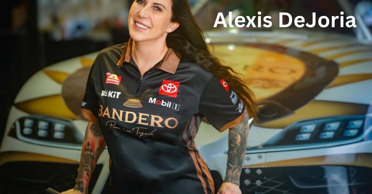 Alexis DeJoria, with tattoos and long dark hair, wearing a black polo shirt with logos, stands in front of a race car.