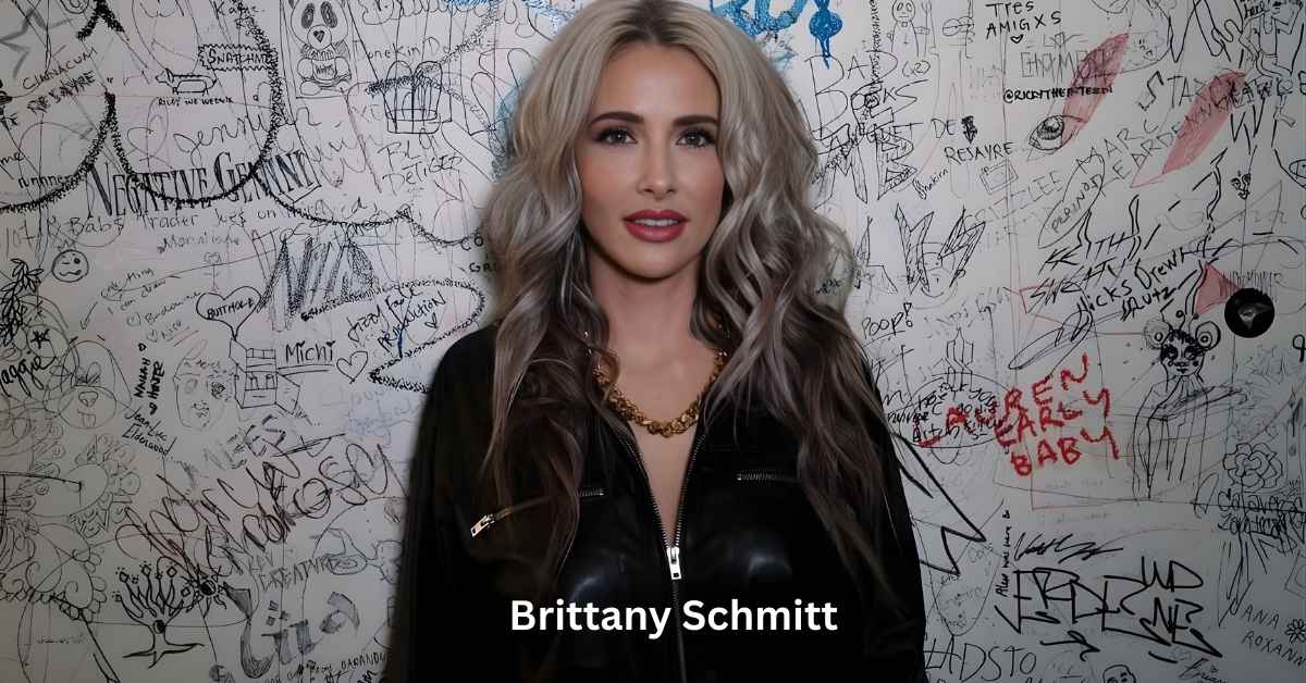 Brittany Schmitt, with long, wavy blonde and grey hair, stands in front of a white wall covered in graffiti and drawings.