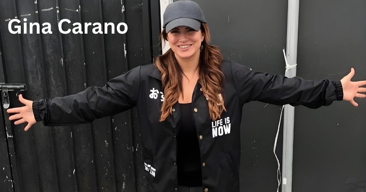 Gina Carano wearing a gray baseball cap and a black jacket with white text that says LIFE IS NOW smiles with arms outstretched in front of a dark, textured background.