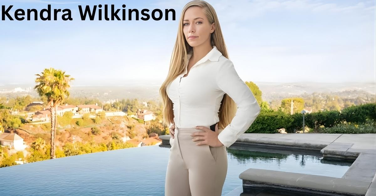 Kendra Wilkinson stands with hands on hips in front of an infinity pool with a city view in the background.