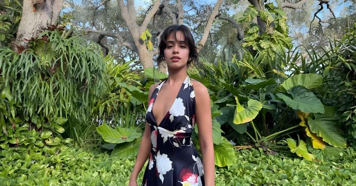 Camila Cabello, in a dark floral dress, stands in a lush garden with large green leaves and trees in the background.