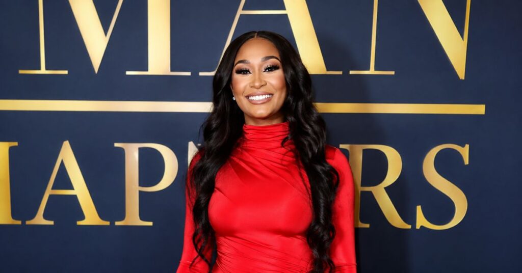 A woman in a bright red high-neck dress smiles for a photo in front of a dark blue backdrop with large gold letters reading “AP AWARDS” and “V I A” at the top.