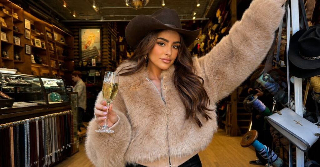 A woman in a boutique wearing a dark wide-brim hat and a light fur jacket holds a champagne flute in a warmly lit store aisle.